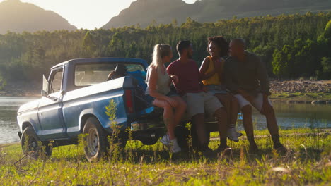 friends with backpacks sitting on tailgate of pick up truck on road trip by lake in countryside