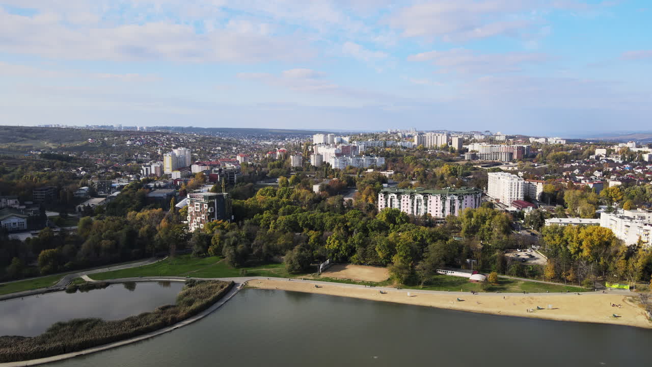 A bright day showcases a beautiful city landscape featuring green parks, residential buildings, and calm waters. People enjoy leisure activities throughout the area