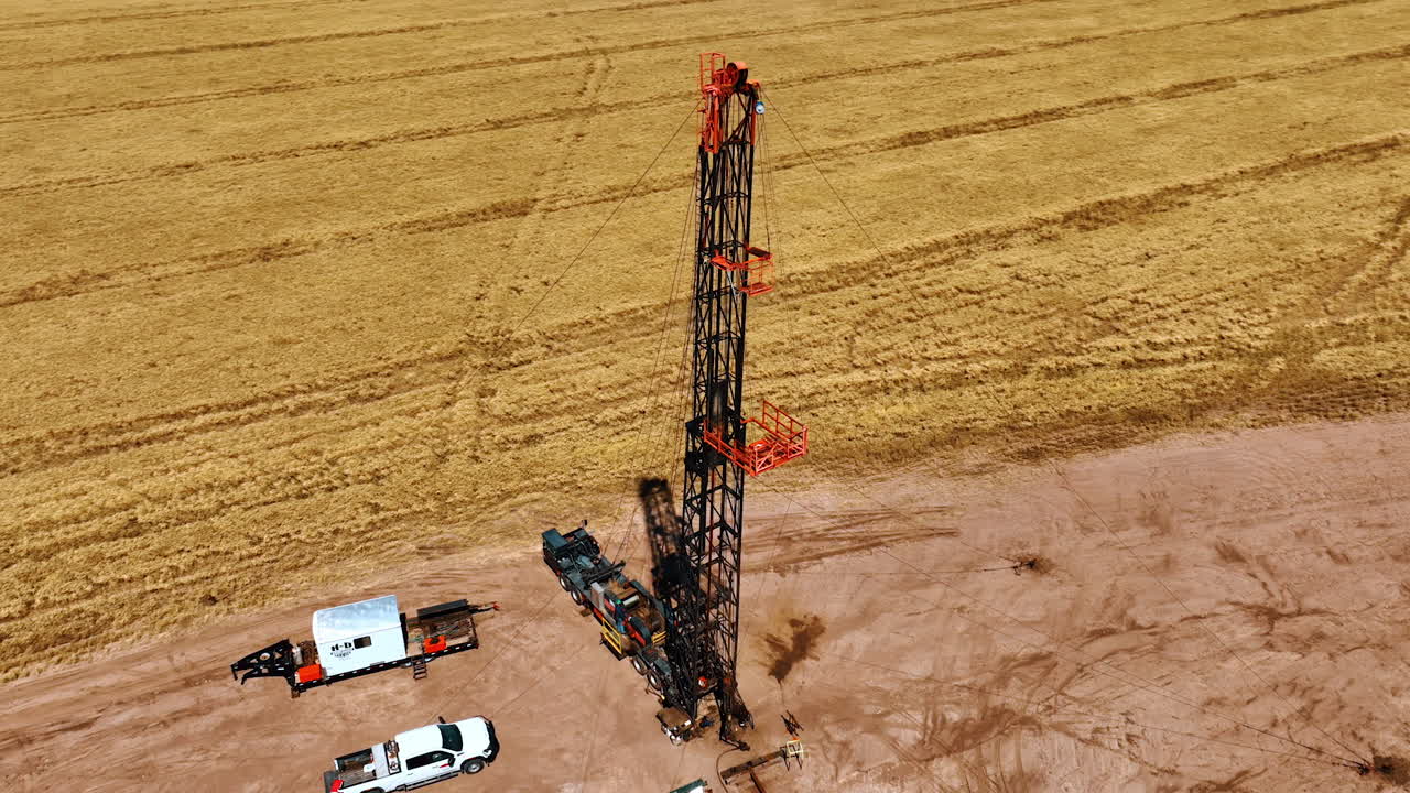 Sunny field with some crops growing on and site for drilling natural resources. Drone footage above the cars and equipment for oil production.