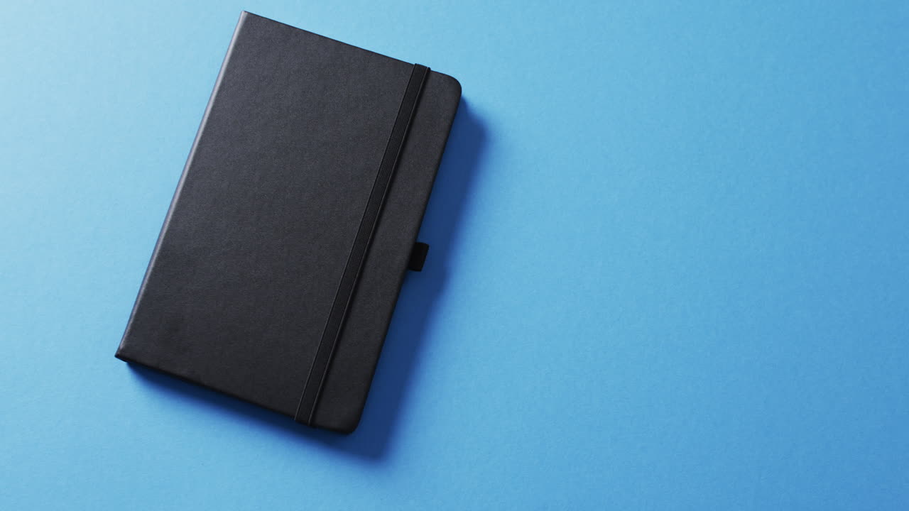 Close up of closed black notebook with copy space on blue background in slow motion