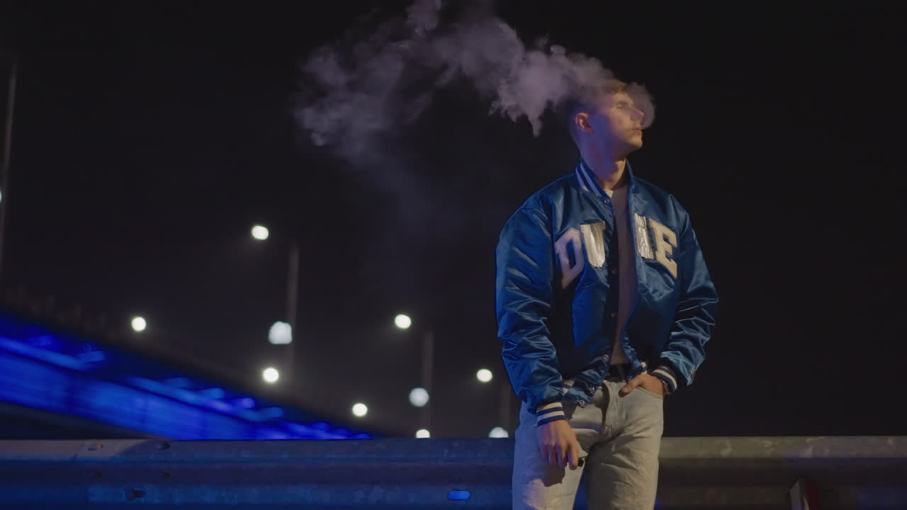 Un hombre blanco exhala humo junto a un puente de noche, con una postura segura y una chaqueta universitaria, la cabeza inclinada mientras el vapor se eleva contra el horizonte iluminado, el contraste cinematográfico y los suaves marcos bokeh evocan a un corredor callejero