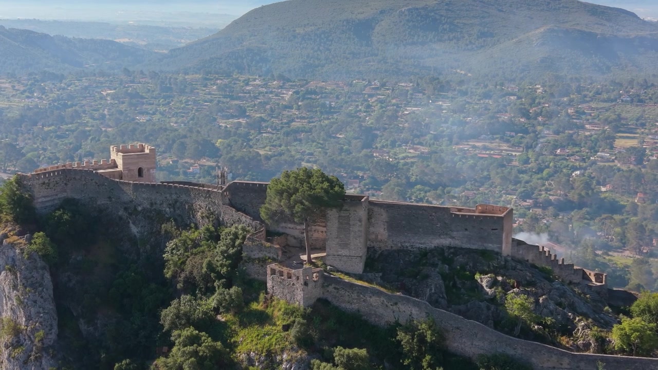 Veduta aerea con drone delle mura difensive del Castello di Xàtiva, arroccato su una scogliera scoscesa sopra valli boscose a Xàtiva, Spagna.