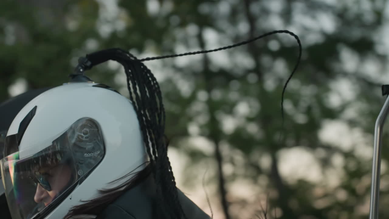 Passenger holds rider closely while traveling on motorcycle near roadside barrier with trees in background, headlight shining brightly as couple enjoys road journey together in warm evening