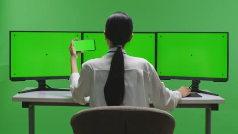 woman using a smartphone in front of a green screen with triple monitor setup