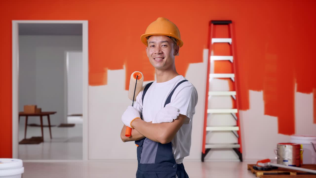 vista lateral de un pintor asiático con casco de seguridad sonriendo y cruzando los brazos mientras está de pie con la pared de pintura en la habitación