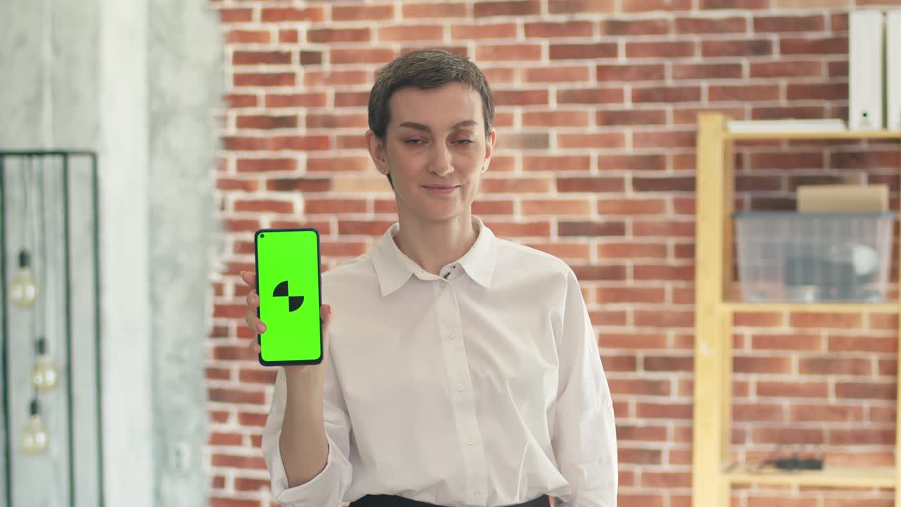 귀여운 사업가,  셔츠, 앱이 있는 드폰을 들고 사무실에 서서 카메라를 쳐다보고 있다.