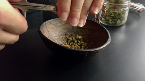 Close-up-of-hands-cutting-weed-in-half-coconut-bowl,-besides-a-jar-full-of-weed