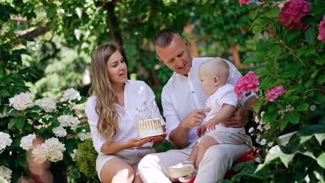 happy parents celebrating the first birthday of their beloved baby boy. mother holds a cake for her little son. beautiful summer garden at backdrop.