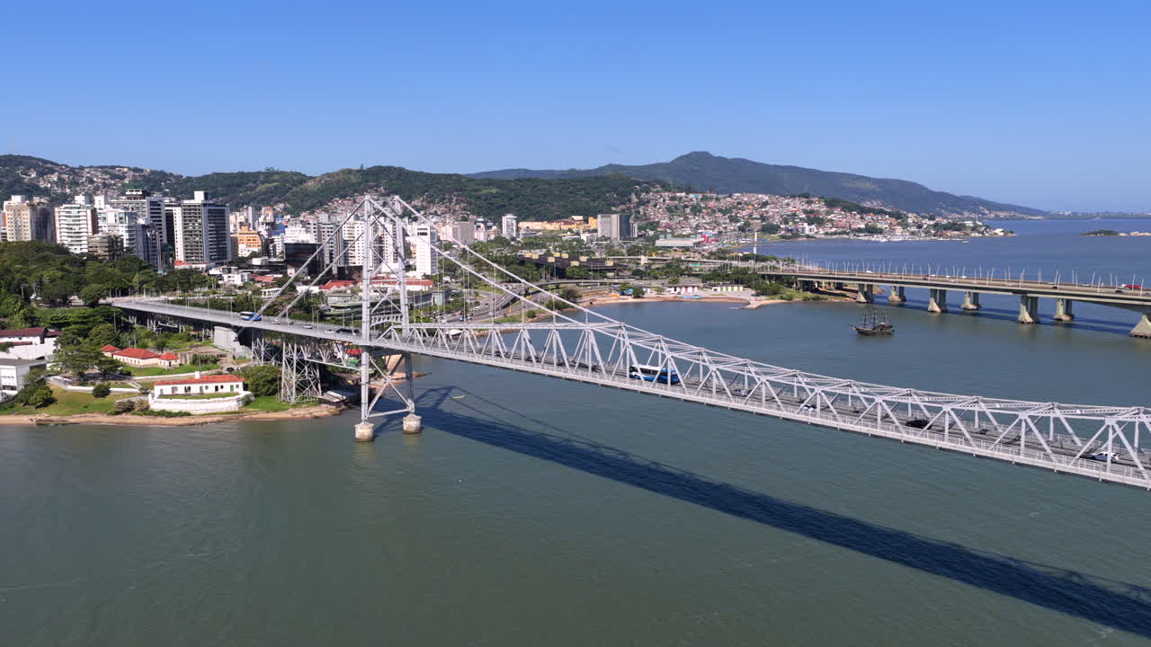 Most Ponte Hercilio Luz ve Florianopolis, Santa Catarina, Brazílie, rozprostírající se nad Baia Norte se spojením Centro a Estreito, vedle mostu Pedro Ivo Campos a městského panoramatu, záběr dronu s oddalováním