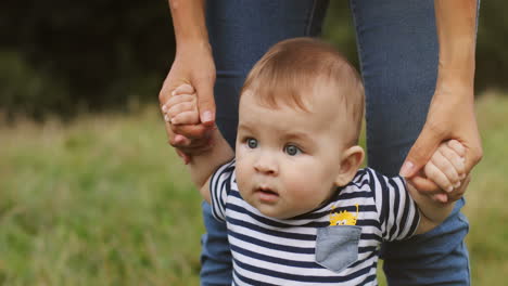 portrait of a cute toddler boy standing and holding his mother's hands outdoors