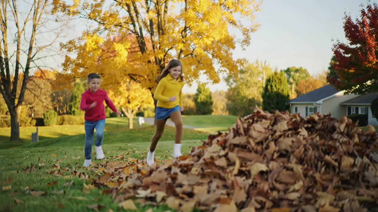 Spotting leaf pile, kids in red hoodie and mustard sweater running, jumping, playing on lawn