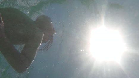 vertical video, young boy swimming underwater, low angle view, camera look up towards sunlight