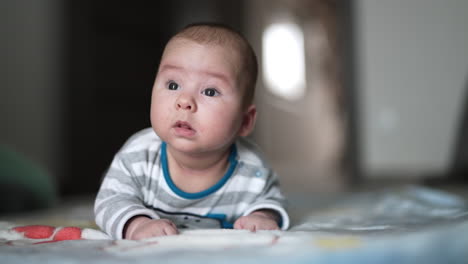 lovely toddler lying on his belly and trying to look up. beautiful boy leaning on his elbows resting on the bed. close up.