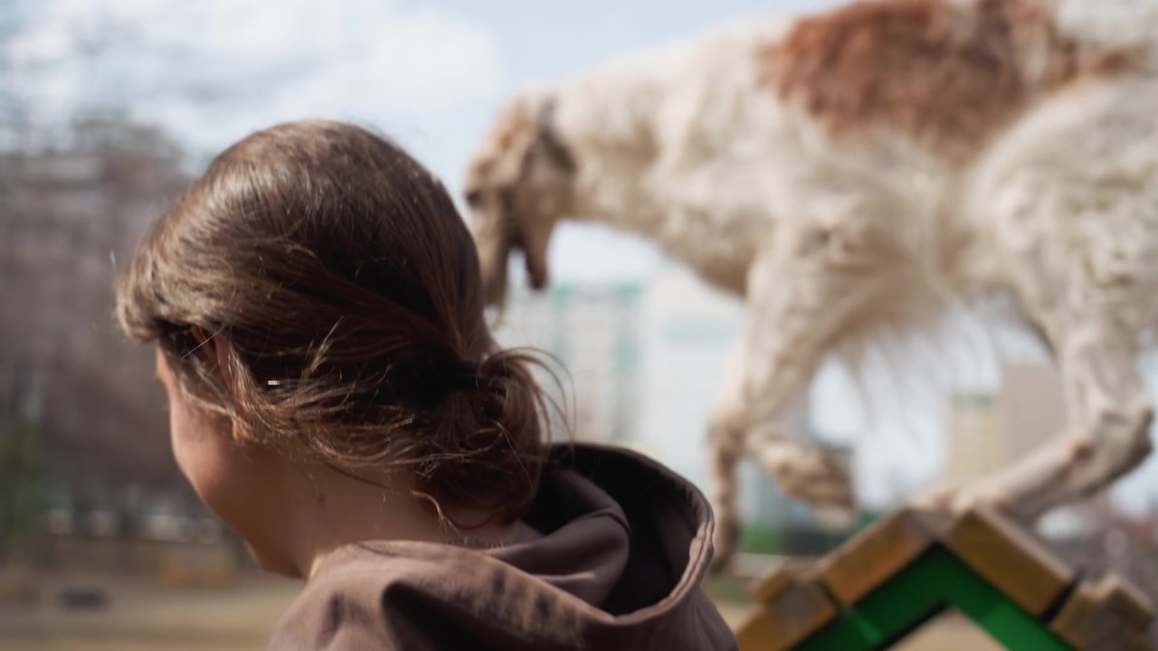 Mujer con sudadera con capucha marrón interactúa con su perro en un parque infantil al aire libre, sosteniendo la correa mientras se mueven entre árboles sin hojas y estructuras de juego verdes. El fondo presenta altos edificios de apartamentos