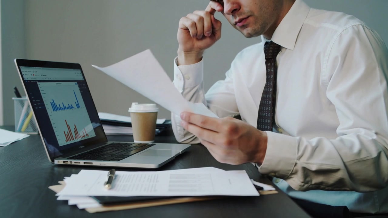 Uomo che sistema, in camicia bianca e cravatta scura, sfoglia rapporti alla scrivania, controllando grafici sul laptop per la riunione