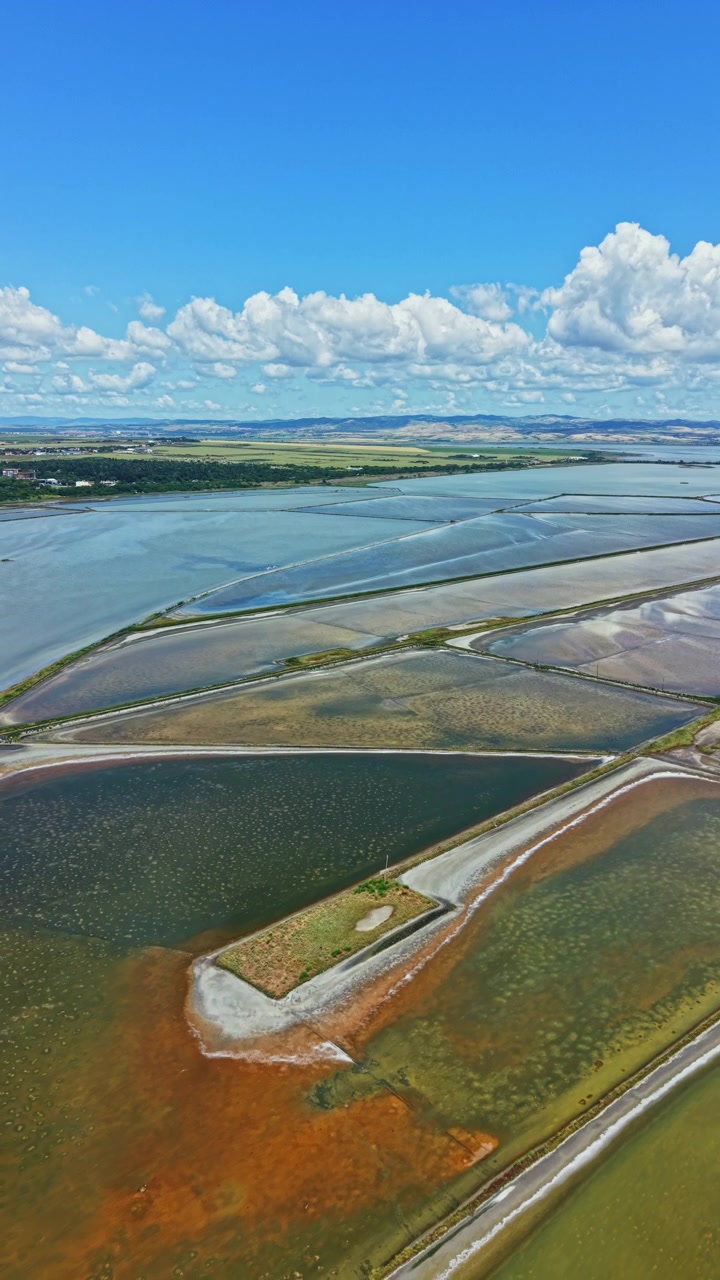 Ampia area di saline e specchi d'acqua vicino a una costa sotto un cielo azzurro e limpido con nuvole sparse.