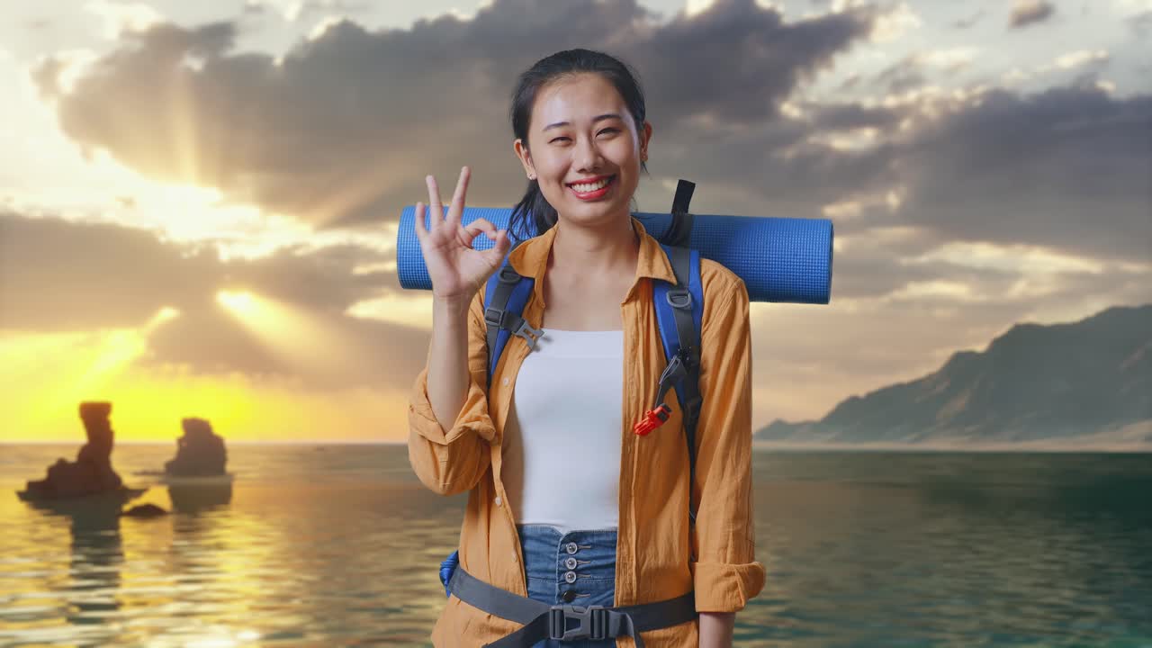 登山用バックパックを背負い、夕暮れ時のビーチに立ってカメラに向かってOKジェスチャーをしながら笑顔を見せるアジア人女性ハイカー
