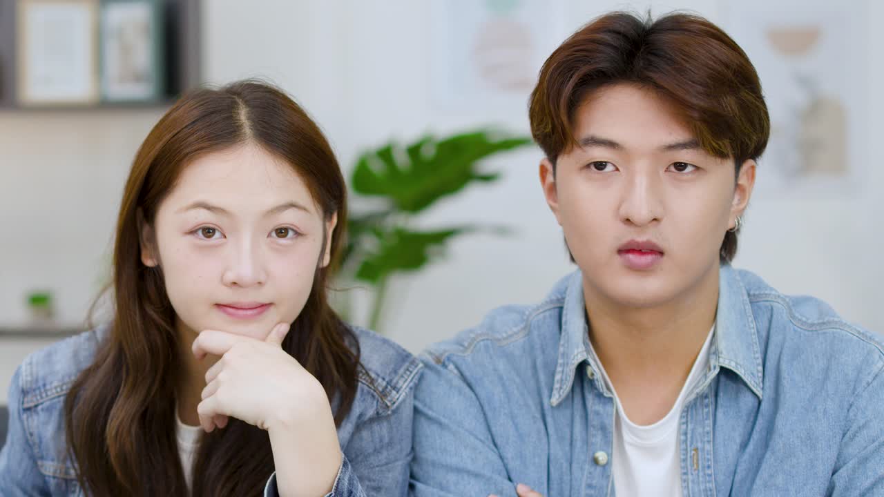 คู่รักชาวเอเชียวัยหนุ่มสาวนั่งอยู่บนโซฟา แสดงสีหน้าท่าทางที่หลากหลายขณะดูโทรทัศน์