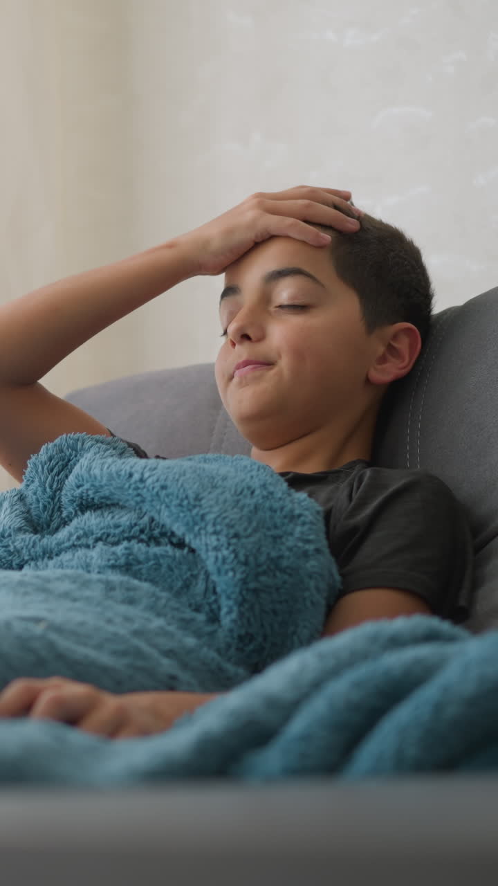 Child suffering from flu cough lies on couch covered with thick blanket, expressing tiredness as he places hand on head and coughs in softly lit room near window with sheer white curtain