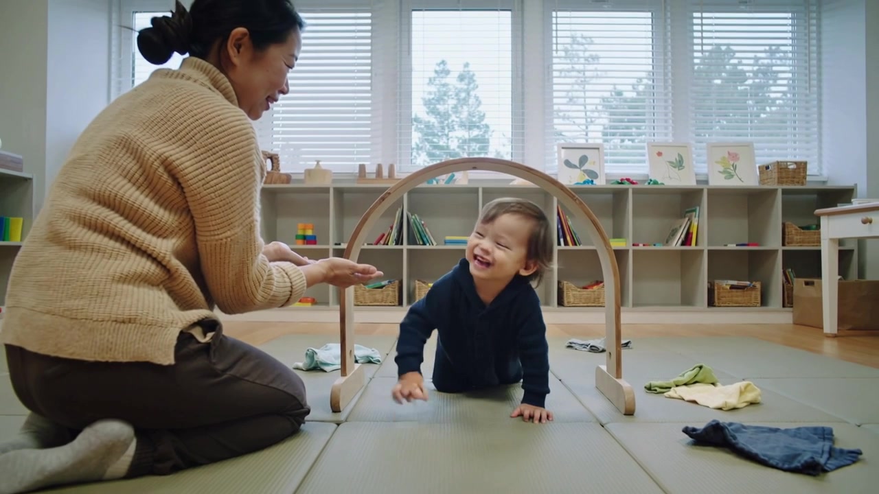 Following caregiver in sweater extending hands, toddler crawling through wooden arch on mat