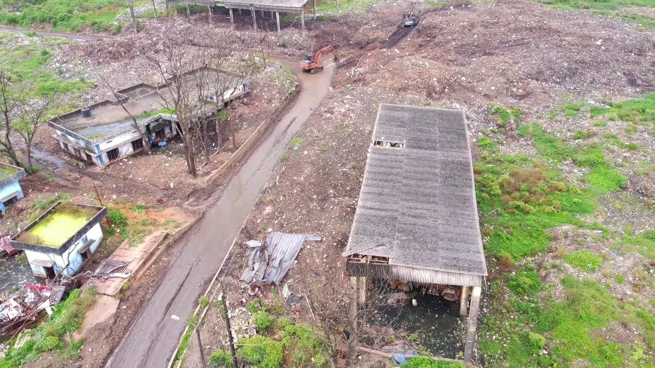Aerial view of a deserted, polluted industrial area with dilapidated buildings, dirt roads, debris, and construction vehicles clearing waste