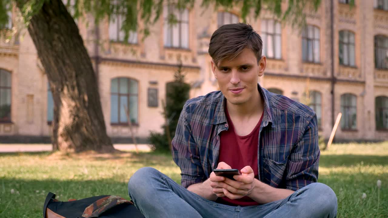젊은 잘생긴 남자가 공원의 잔디 위에 앉아 스마트폰을 하고 있습니다. 휴식 개념, 의사소통 개념, 배경에 기반을 두고 있습니다.