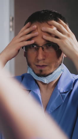 slow motion: young doctor in blue uniform with glasses takes off white surgical mask preparing to work near large mirror in public bathroom closeup