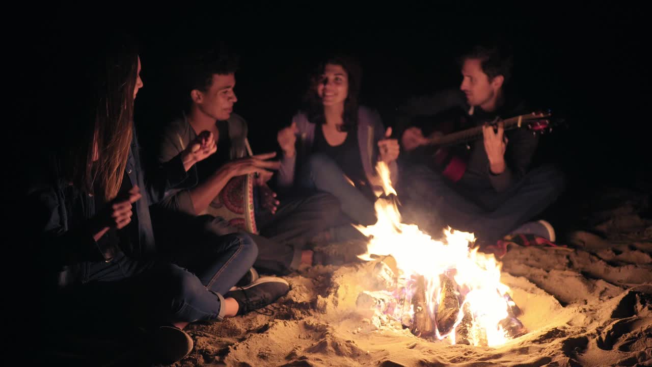 dos mujeres jóvenes y dos hombres sentados junto a la hoguera tarde en la noche y cantando canciones, tocando la guitarra y la percusión