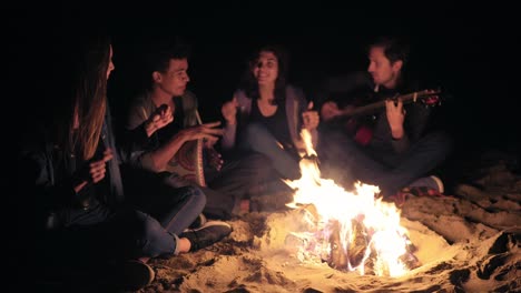 two young women and two men sitting by the bonfire late at night and singing songs, playing guitar and percussion