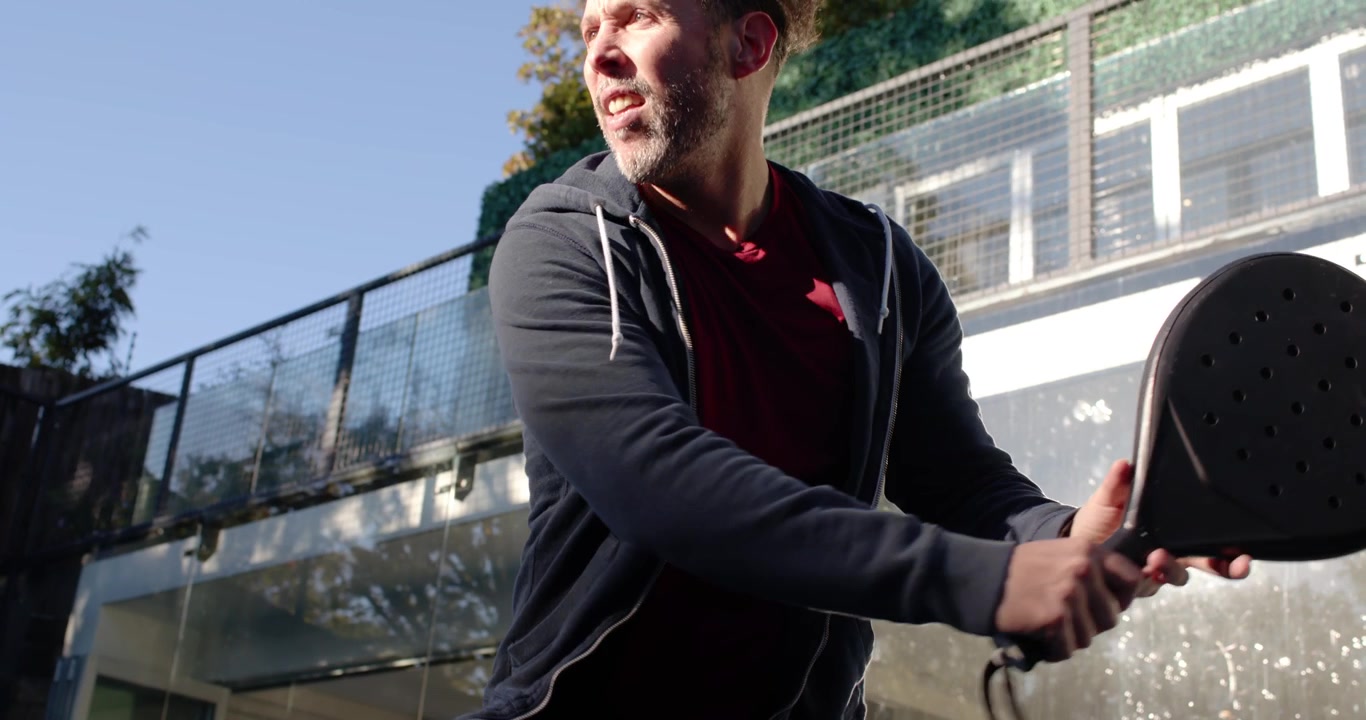 Diverse men reacting to incoming ball on padel court, player shifting stance and swinging paddle