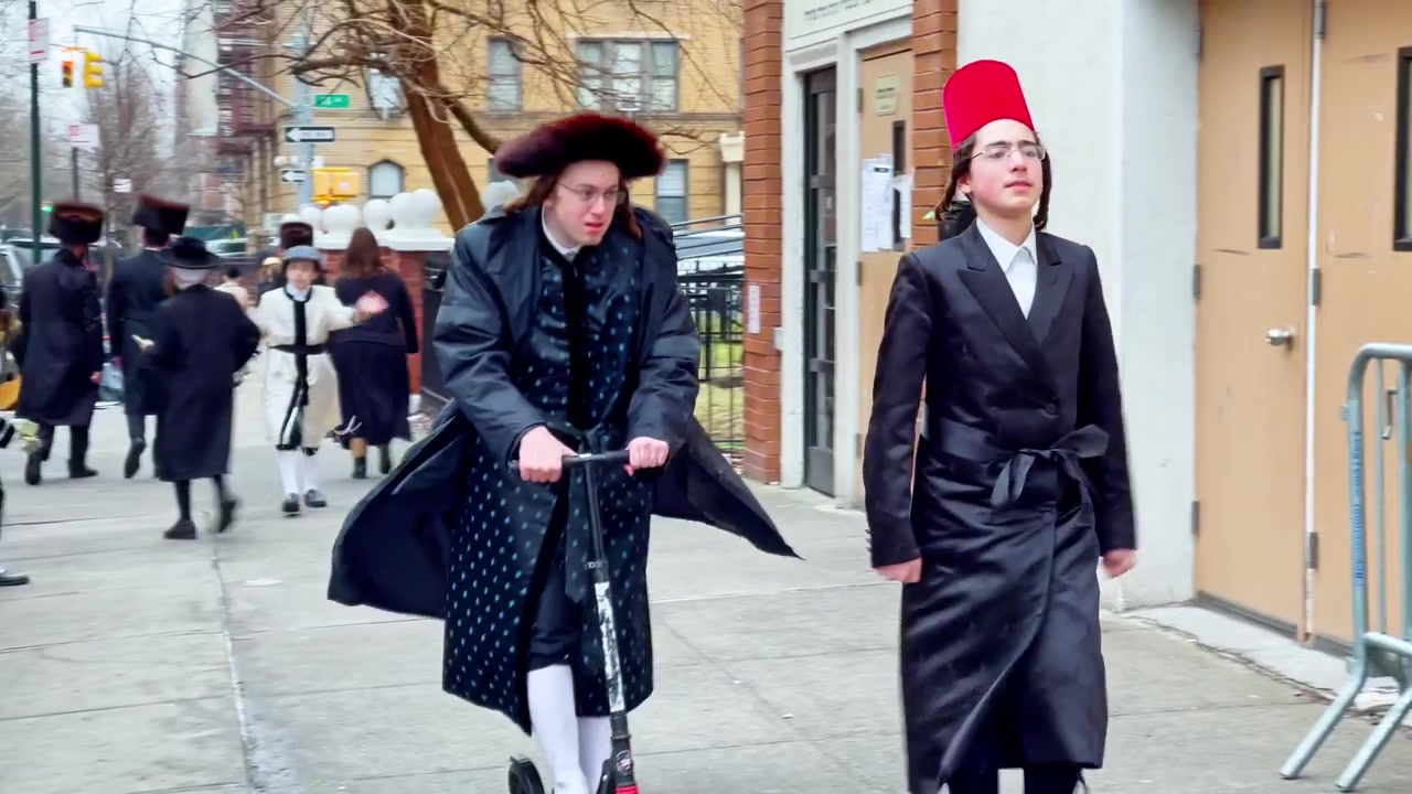 New York, USA, 9. marts 2022: Folk i traditionelle hatte går på fortovet i Brooklyn. To unge mennesker i sorte frakker og hatte bevæger sig langs fortovet i Brooklyn, den ene kører på et løbehjul.