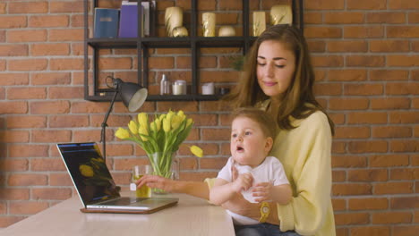 side view of woman sitting on kitchen counter working on laptop while holding baby on her lap