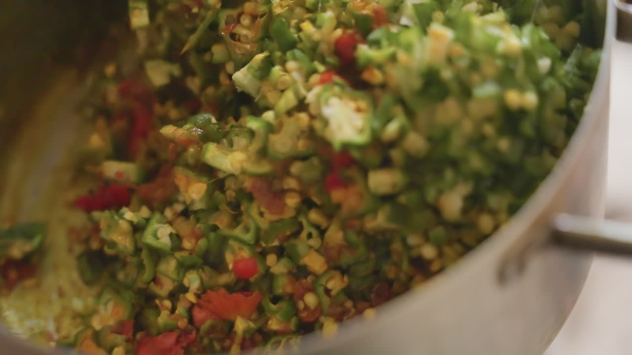 Primo piano di una mano nera che mescola verdure in una pentola d'acciaio inossidabile, broccoli tritati, pomodorini e mais conditi con olio ed erbe aromatiche, movimento di un cucchiaio blu, luce calda della cucina, cuoco casalingo che prepara una cena sana.