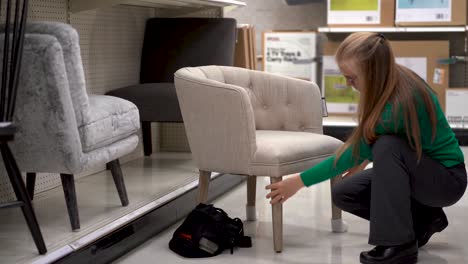 squatting down, a blonde woman scrutinizes an upholstered, modern looking chair in a store display