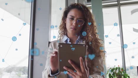 una mujer de negocios biracial sentada en un escritorio y usando una tableta en interiores de oficina modernos