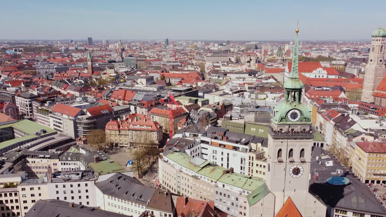 Munich’s Marienplatz reveals the Neues Rathaus clock tower rising above red-tiled roofs, bustling streets, and Gothic spires blending medieval charm with modern city life, drone establishing shot