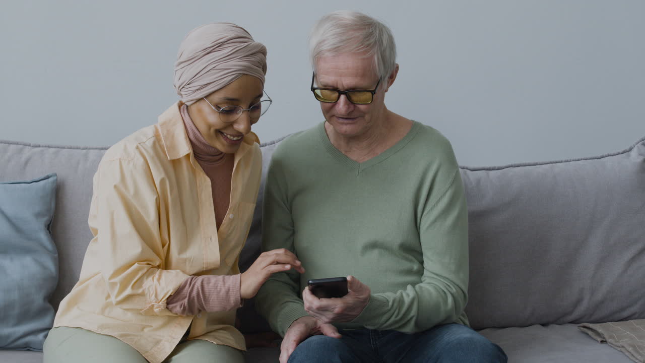 Smiling Middle Aged Arabic Woman Helping A Senior Man To Sync Smartphone To Robot Vacuum Cleaner While Sitting On Sofa At Home 1