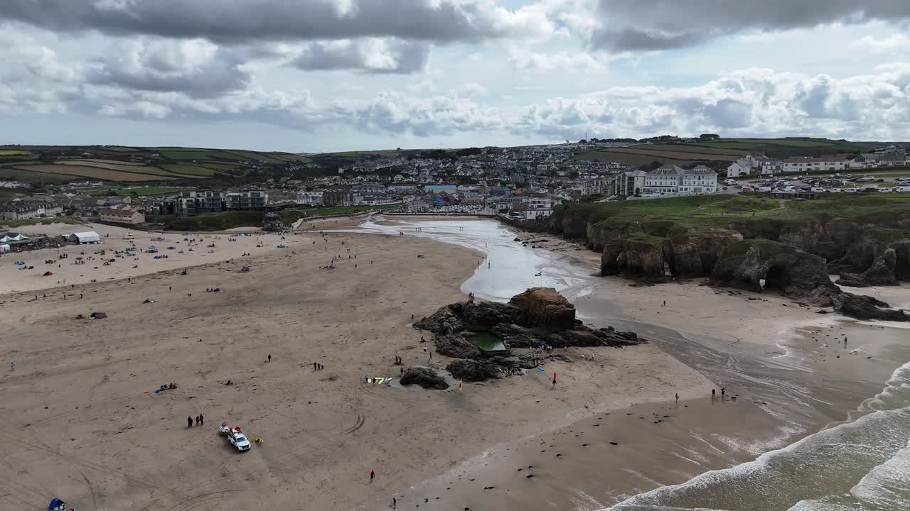 Luftfoto med drone af en bred sandstrand i Devon på Englands sydkyst, der viser rullende bølger, mennesker langs kystlinjen, klippeklipper og en kystby under en skyet himmel