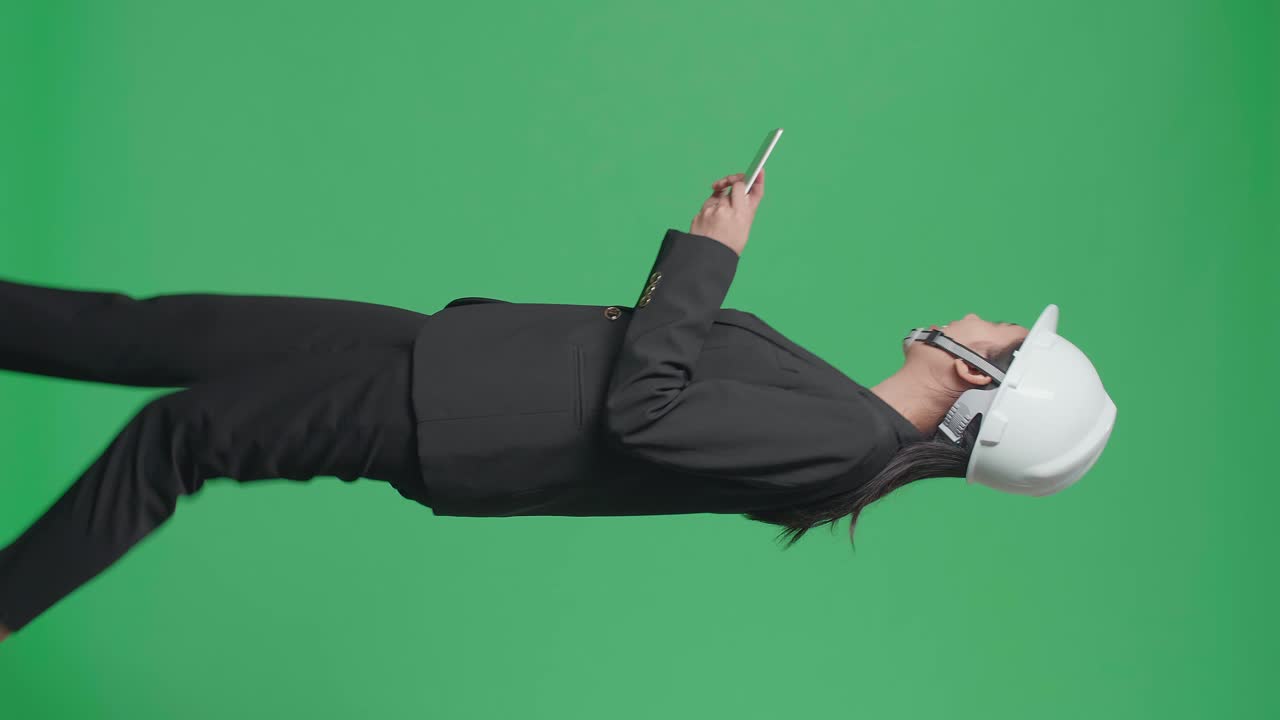Side View Of Smiling Asian Female Engineer Looking Around And Shaking Her Head While Using Tablet And Walking In The Green Screen Studio