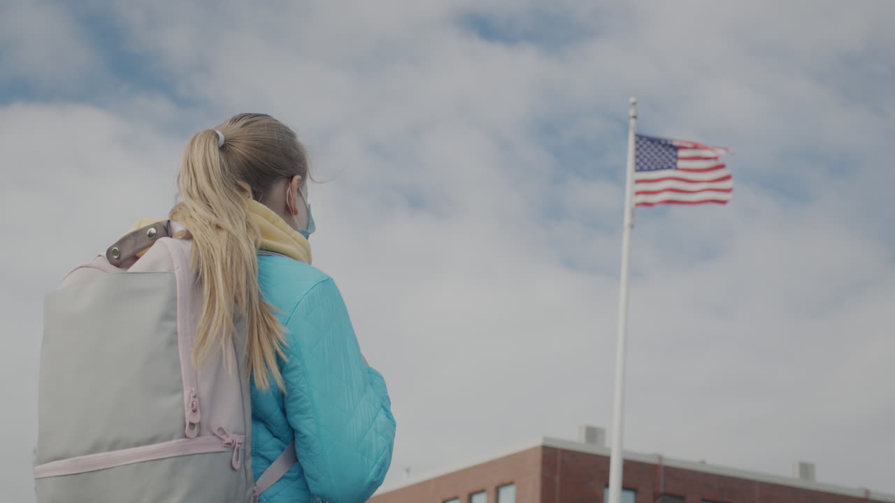 vista trasera: una colegiala con una máscara protectora se encuentra contra el fondo de una escuela y una bandera estadounidense.