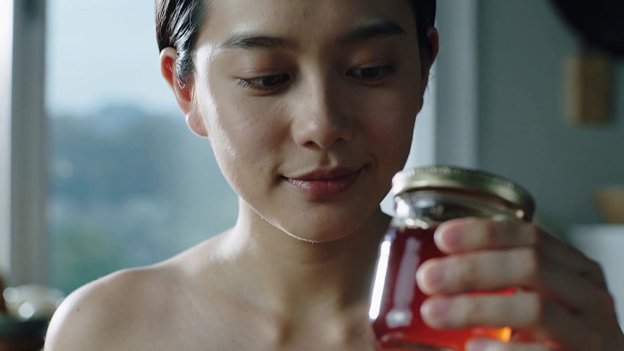 Primo piano di una donna asiatica che solleva un barattolo all'altezza degli occhi in cucina, studiando un liquido ambrato.