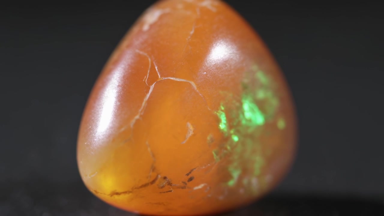 Kamera fährt zurück und enthüllt einen dreieckigen orangefarbenen Cabochon auf einer dunklen Plattform, der einen grünen Schimmer aufweist.