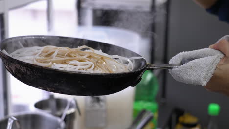 Chef-pours-ladle-of-dark-sauce-over-rice-noodles-in-skillet,-pan-filled-with-steaming-rice-sticks-get-doused-in-soy-sauce,-slow-motion-close-up-4K