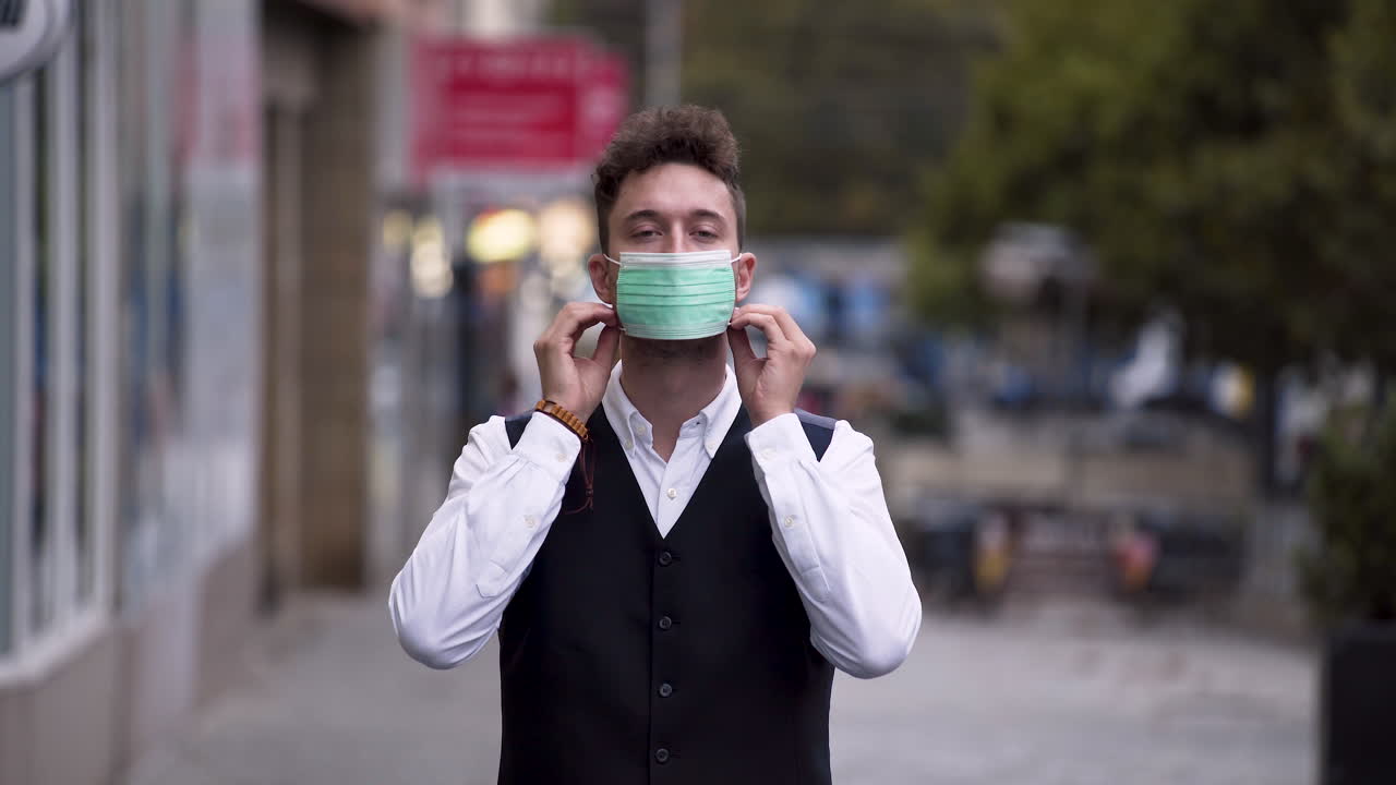 An elegant young man wearing a black waistcoat and a white shirt putting on a green protective Covid-19 mask, standing on a busy street, looking straight into the camera, static close up 4k shot