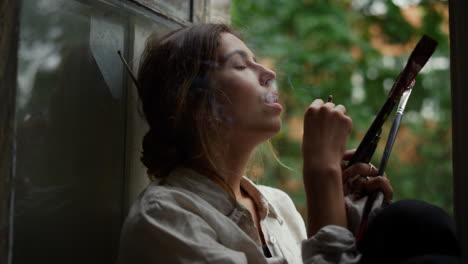 tired painter smoking on windowsill. young female artist resting in art studio.