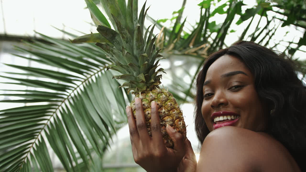Young girl with a bare top african american mixed race holds a pineapple in her hands and smiles at