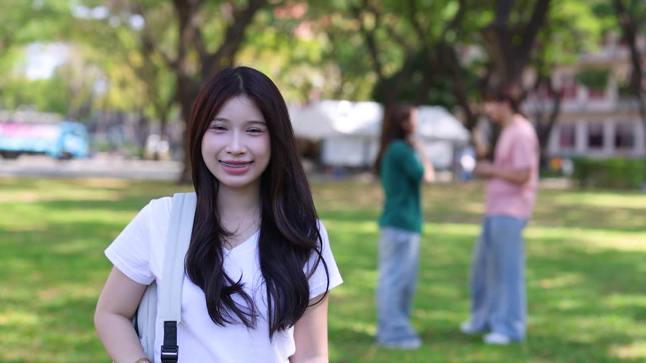 หญิงเอเชียอายุน้อยสวมเสื้อเชิ้ตสีขาว ยิ้มขณะสะพายกระเป๋าเป้กลางแจ้ง
