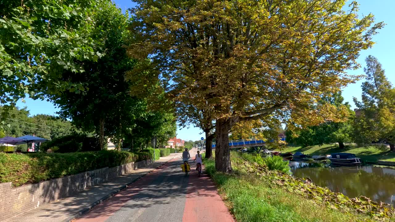 Two cyclists travel on a tree-lined bike lane beside a canal in bright summer daylight