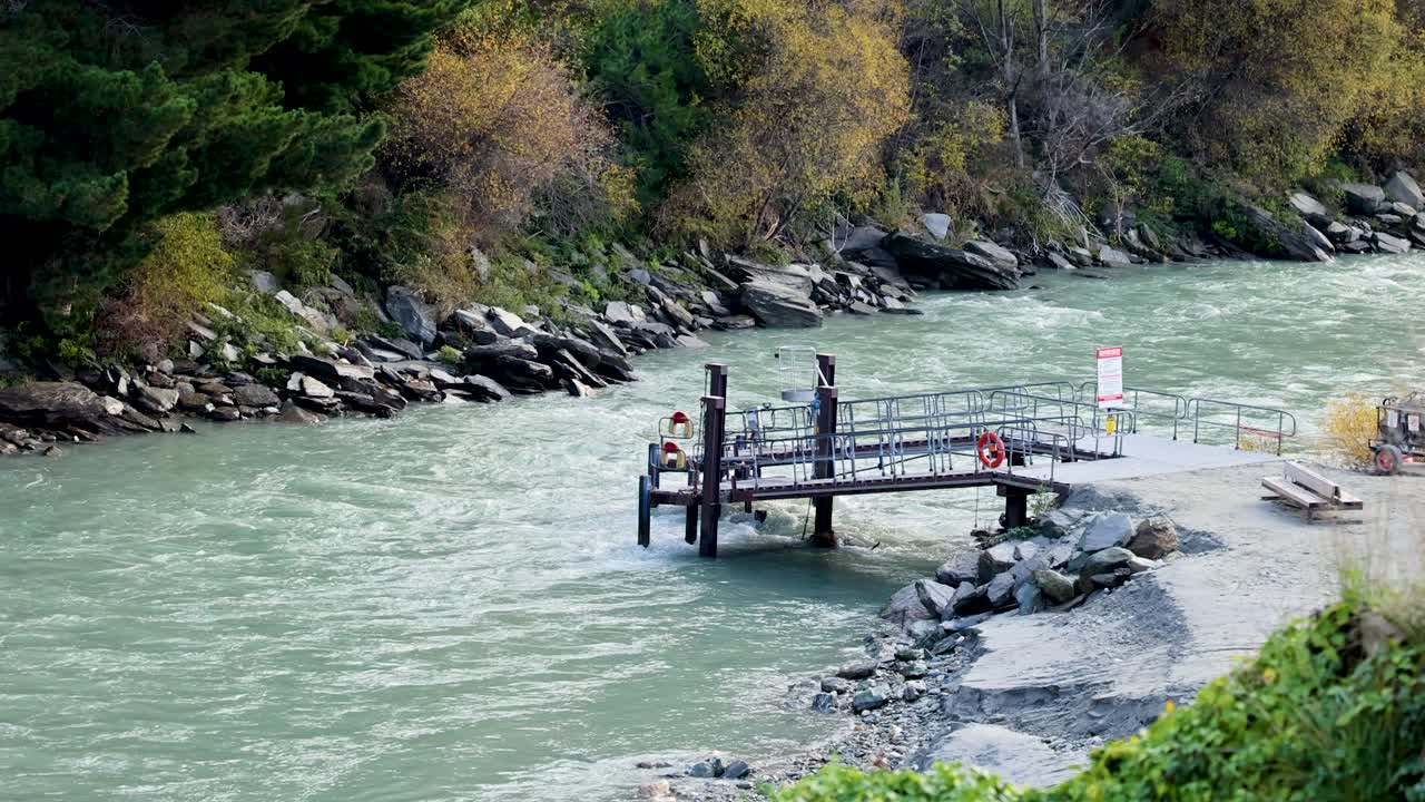 A tranquil river flows past a wooden dock surrounded by lush greenery and autumn foliage under soft, natural lighting