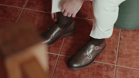close up of man in light trousers fixing socks while wearing formal brown monk strap shoes